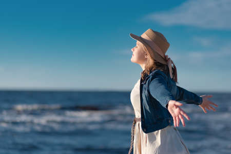 A woman with blond hair in a straw hat, a beige dress and a denim jacket stands on the background of the sea and enjoys the sun. The concept of peace of mind and happiness.の写真素材