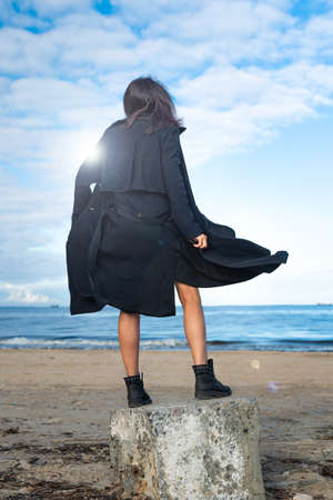 young woman in a black cloak developing in the wind and boots, stands on a stone by the sea.の写真素材