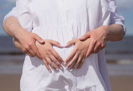 pregnant woman on the beach in a white dress, holds her hands on her stomach, folded them in the shape of a heart.の写真素材