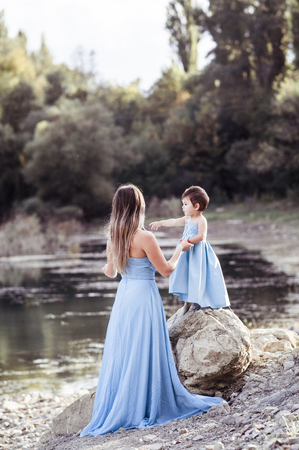 Beautiful mother with small daughter in blue indentical dresses standing on stone near riverの写真素材