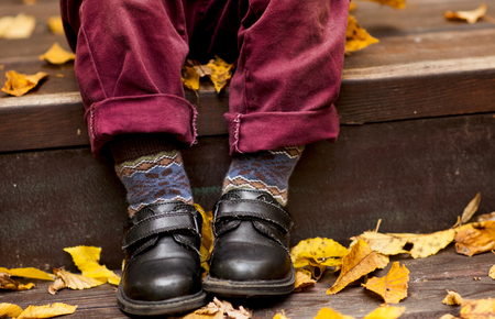 Child boy  dressed in crimson trousers and black shoes with colored socks, seats on wooden stairs in...