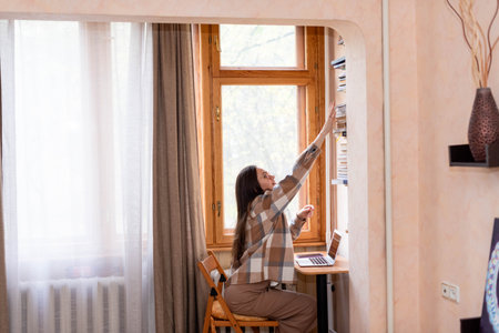 Freelancer girl takes books from the shelf, works at a laptop near the windowの写真素材