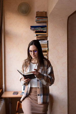 A woman in glasses is taking notes with a pen in a notebook near a table with a laptopの写真素材