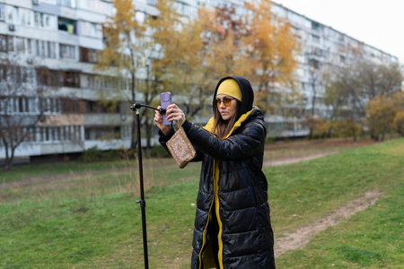 Female blogger in orange glasses preparing to record video on phone with tripod in open areaの写真素材