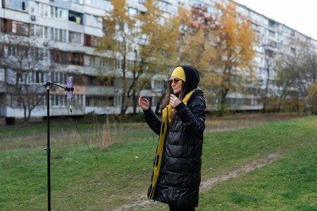 Female in orange glasses preparing to record video on phone with tripod in open areaの写真素材