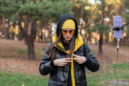 Female blogger in orange glasses preparing to record video on phone with tripod in open area