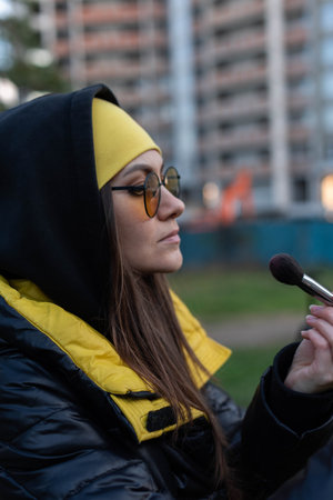A girl in orange glasses fixes her makeup on the street - uses a mirror and a brushの写真素材