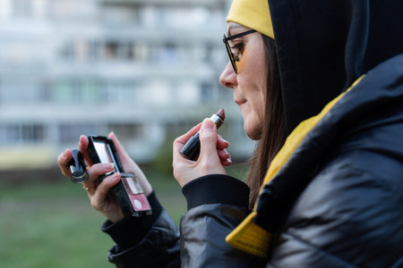 A girl in orange glasses fixes her makeup on the street - uses a mirror and a lipstickの写真素材