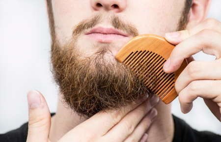 Closeup of a young man styling his long beard with a comb while standing alone in a studio against a...