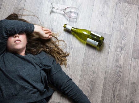 Drunk alcoholic young girl lying on the floor feeling sick by drinking red wine, empty glass and bottleの写真素材