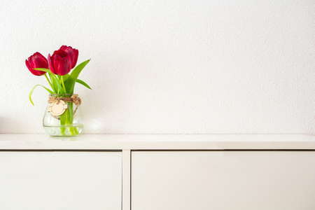 Red tulips in tall glass jar on white table against white wall modern bright design, spring concept...