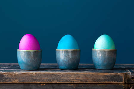 Colorful bright Easter eggs hand painted in egg cups on a blue background on wooden shelf Holiday...