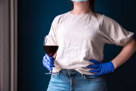 Woman wearing protective mask and blue gloves in her home and drinking red wine, self isolated for...