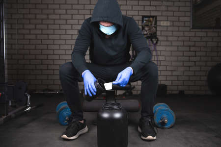 Young healthy man in gym preparing Protein Shake Before Workout for building muscle faster in black workout clothes with protective mask and gloves for Coronavirus, Covid-19の写真素材