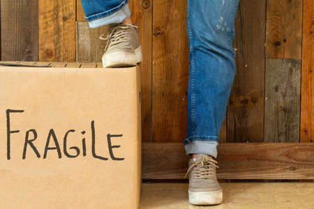 Moving box with the word Fragile near wooden wall, two legs with jeans and sneakers in retro room...