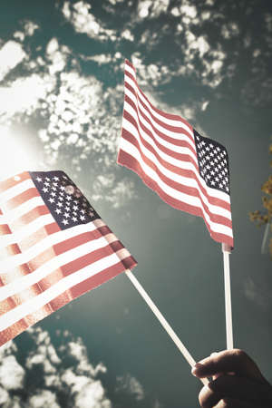 Hand holding two american flags on the blue sky with sunlight background, waving flag for United States of America close-upの写真素材