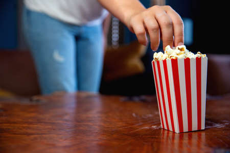 Beautiful young woman sitting on a living room couch, watching television with a red and white striped cardboard box with popcorn retro popcorn boxの写真素材