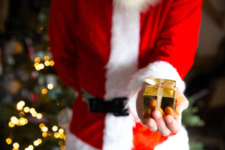 Man in Santa Claus costume near Christmas tree holding a golden gift box, Christmas present background with bokeh lightsの写真素材