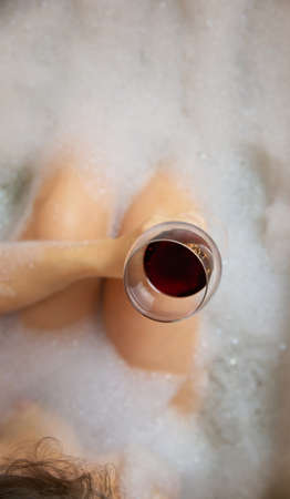 Young womans hand holding wine glass in bathtub, drinking red wine and lying in bathtub with bubble foam, top view, relaxtion and spa conceptの写真素材