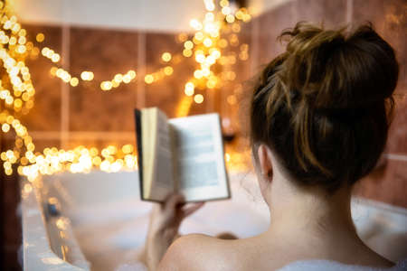 Young beautiful woman reading a book and drinking red wine in the bathtub with bubble foam, decorated with colorful lights, relaxing and spa concept close-upの写真素材