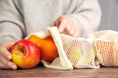 Female hand with environmentally friendly net bag for shopping groceries with Fresh fruit, Apples and oranges, eco friendly net on wooden tableの写真素材