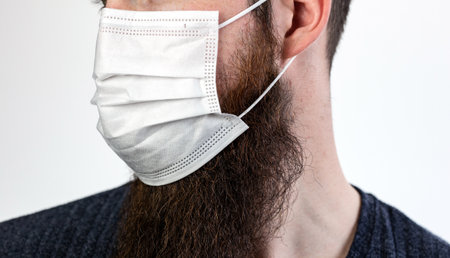 Caucasian young bearded adult man wearing a protective or surgical mask to avoid pollution and contagious virus and diseases isolated on white backgroundの写真素材
