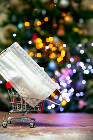 Protective safety mask in iron shopping basket near Christmas tree, Covid-19, coronavirus, holiday concept with copy space and bokeh lights backgroundの写真素材