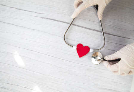 Red heart and white protective medical gloves holds a stethoscope on a white wooden background. Concept visit to the doctor, virus, treatment, diagnosis. Banner. Flat lay, top view copy spaceの写真素材