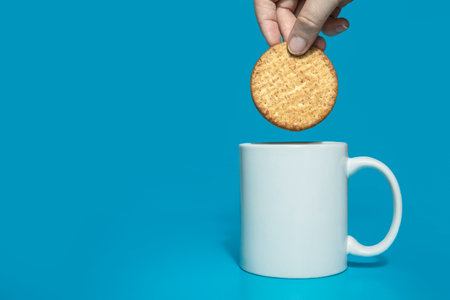 Female hand dipping normal healthy grain cookies in white mug of milk on blue background, copy space.Dairy,drink concept tasty snackの写真素材