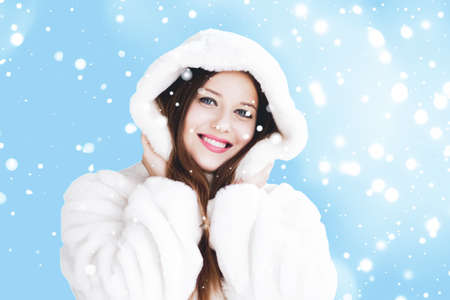 Christmas and winter holiday portrait of young woman in white hooded fur coat, snow on blue...