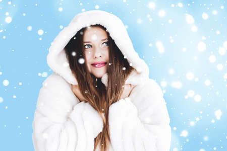 Christmas and winter holiday portrait of young woman in white hooded fur coat, snow on blue background, fashion and lifestyle campaignの写真素材