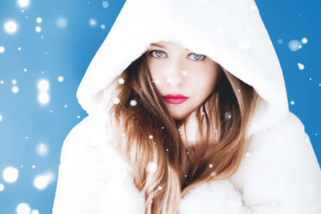 Happy Christmas and winter holiday portrait of young woman in white hooded fur coat, snow on blue...