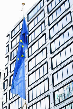 European Union flag waving in front of modern corporate office building, symbol of EU Parliament,...