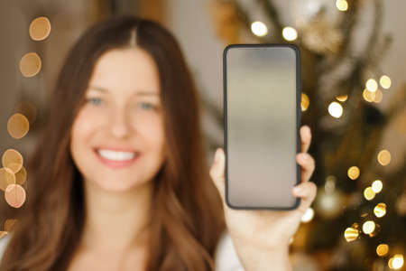 Christmas phone call and holiday greeting concept. Happy smiling woman showing screen of mobile smartphone, xmas decoration on backgroundの写真素材