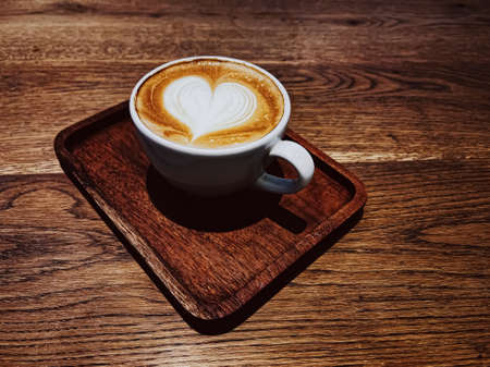 Coffee break and romantic mood concept. Cup of caramel cappuccino with heart shaped foam art made of lactose-free milk, served on wooden table in cafeteriaの写真素材