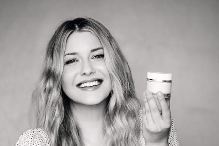 Happy young woman with wavy blonde hair holding a face cream moisturiser jar, promoting skincare and...