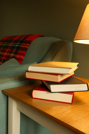 Stack of books sitting on a rustic wooden table, bathed in the soft glow of a lamp, creating a warm and inviting setting perfect for reading and gaining knowledge in the comfort of homeの写真素材