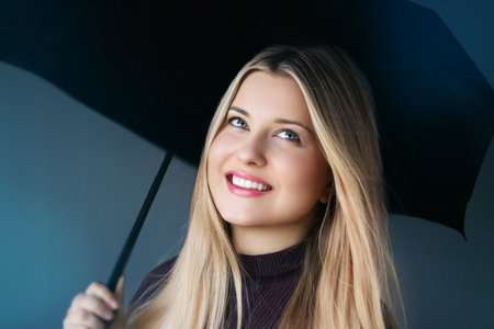 Young woman smiling, holding black umbrella, feeling positive and hopeful, looking up with bright blue eyes, finding joy and optimismの写真素材