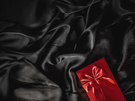 Red gift box with a ribbon bow offering a luxurious surprise on black silk backdrop, symbolizing celebration and eleganceの写真素材