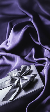 Silver gift box with shiny bow resting on luxurious purple silk fabric, creating an elegant background for celebrationsの写真素材