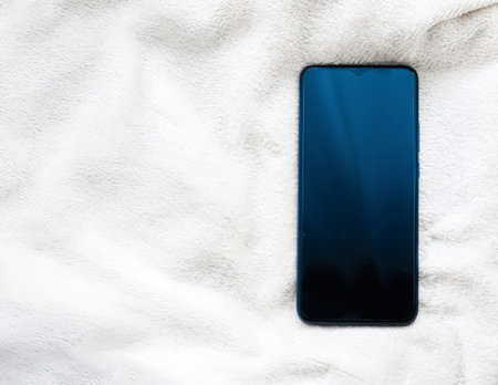 Modern mobile phone with a dark screen lying on a textured white fabric, representing digital comfort and relaxationの写真素材