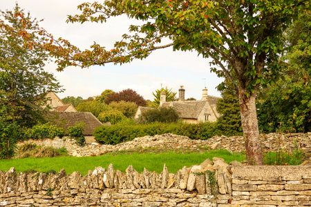 Traditional English village scene featuring rustic stone cottages, dry stone walls, green fields,...