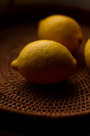 Fresh yellow lemons resting on a textured woven wicker tray, creating a dark moody still life arrangement with natural lightの写真素材