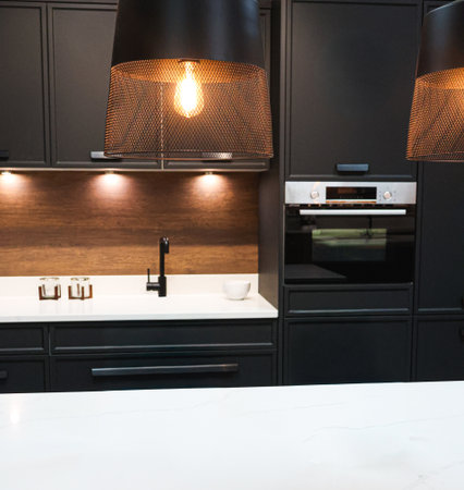 Modern kitchen interior showcasing dark cabinets with an integrated oven, white countertop, warm wood backsplash, and a stylish pendant lightの写真素材
