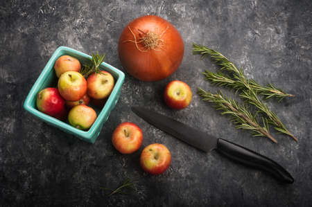 Overhead view of a kitchen scene showing crated mini apples, onion and rosemary along with a knife...