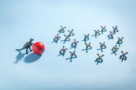 A strong dinosaur pushes a red plastic ball toward a group of vintage jacks pieces. A conceptual photo illustrating strength, bullying, fighting or competition.の写真素材