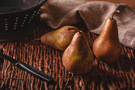 Still life scene of three pears, a paring knife, colander and towel on a woven twig background.の写真素材