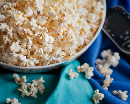 A spilled bowl of home made popped, buttered popcorn on a blue blanket with a remote control nearby to kick off movie night.の写真素材