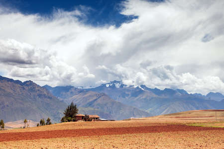 White clouds above the mountains, in the foreground a plowed agricultural field. The land is red, behind it stands a house, behind a house mountains in the clouds.の写真素材