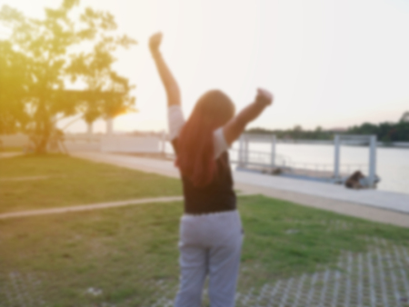 Back view portrait of a  teenage woman raising arms at warm sun at sunrise on the river, defocus blurred for backgroundの写真素材